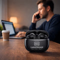 Zwarte EGS Eargearshop draadloze oordopjes in open oplaadcase op een houten bureau met laptop en werkende man op de achtergrond. 🎧💻
